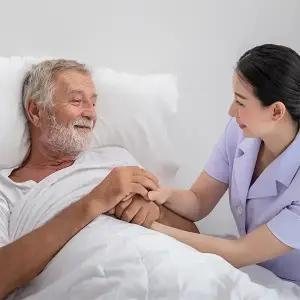 Nurse holding hands and smiling with an elderly man lying in-bed